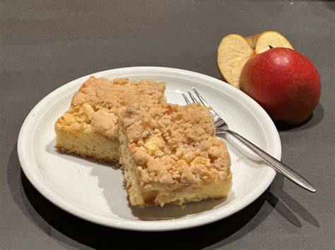 Angeschnittener Apfelstreuselkuchen auf einem Holzteller, dazu eine Kugel Vanilleeis und Sahne