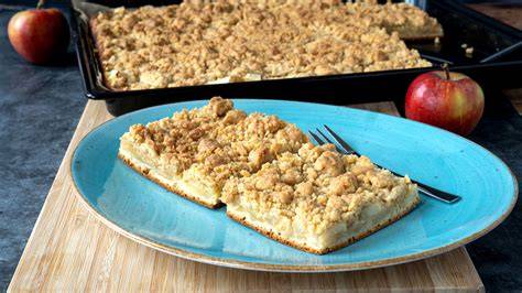 Apfelkuchen mit Streusel angerichtet auf einem Blech, im Hintergrund Äpfel