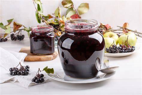 Gläser mit Aronia-Pflaumen-Marmelade auf Holzbrett, daneben frische Zwetschgen und Aroniabeeren