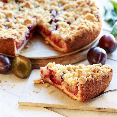 Pflaumenkuchen ohne Streusel auf rustikalem Brett, halbierte Pflaumen und Messbecher mit Mehl