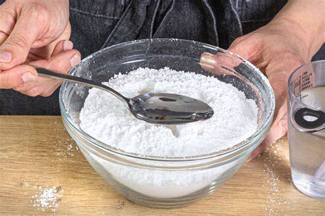 Schüssel mit gesiebtem Puderzucker und Messlöffel Wasser