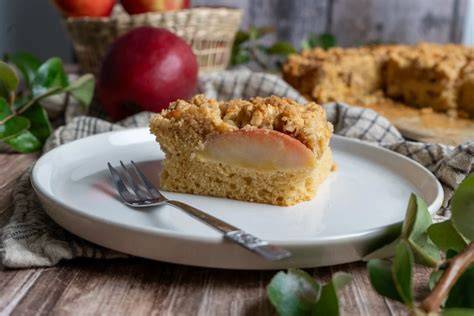 Stück Apfelkuchen mit Streuseln auf Holzbrett, daneben Äpfel und ein Glas Milch