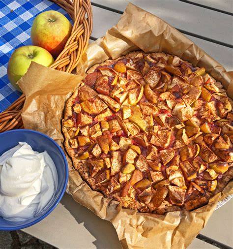rustikaler Apfelkuchen auf Holzbrett, daneben Äpfel und Zimt